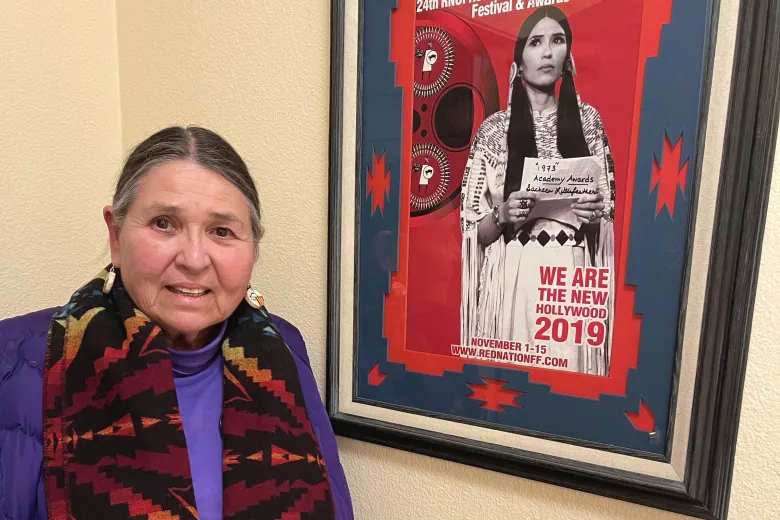 A person with salt and pepper hair slicked back, wearing a colorful scarf around their shoulders, standing next to a poster with a picture of their younger self. 