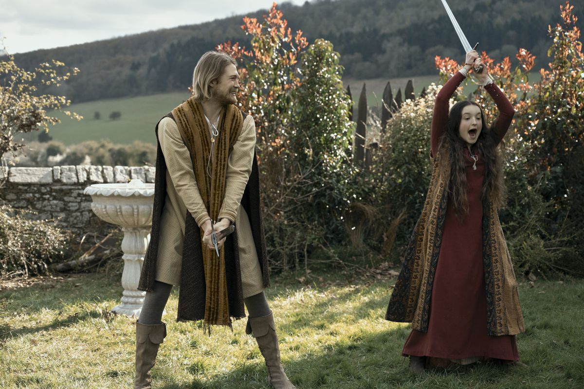 a young teenage girl in medieval clothing raises a sword above her head;  next to her, a blond man smiles and looks at her affectionately 