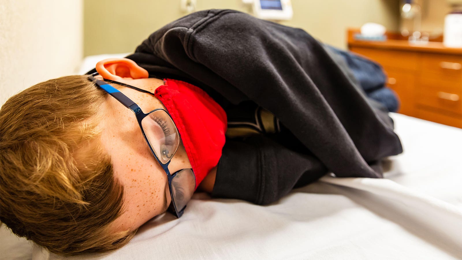 A photo of a little boy wearing a protective mask and wrapped up in a jacket and laying on an examination table.