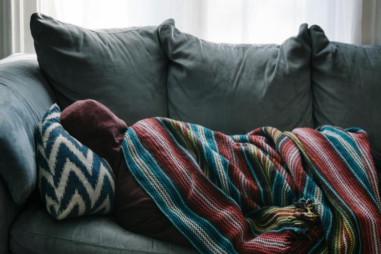 The person is lying on the couch in a hoodie under a blanket.