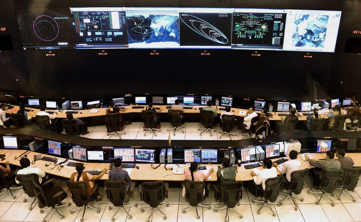 In a kind of theater, scientists sit at rows of desks with computers.  Large monitors stand at the front of the room, which they all face.