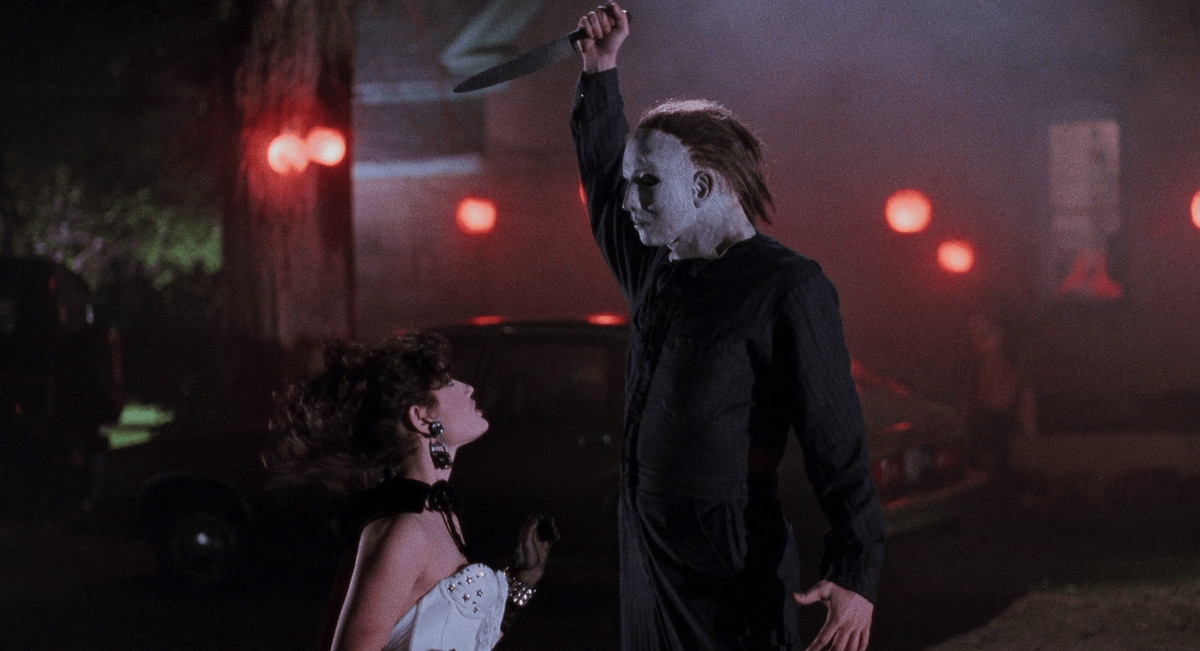 A shaggy Michael Myers stiffly stretches his arm over his head in Halloween 5: The Revenge of Michael Myers, ready to stab a kneeling woman in a white dress.