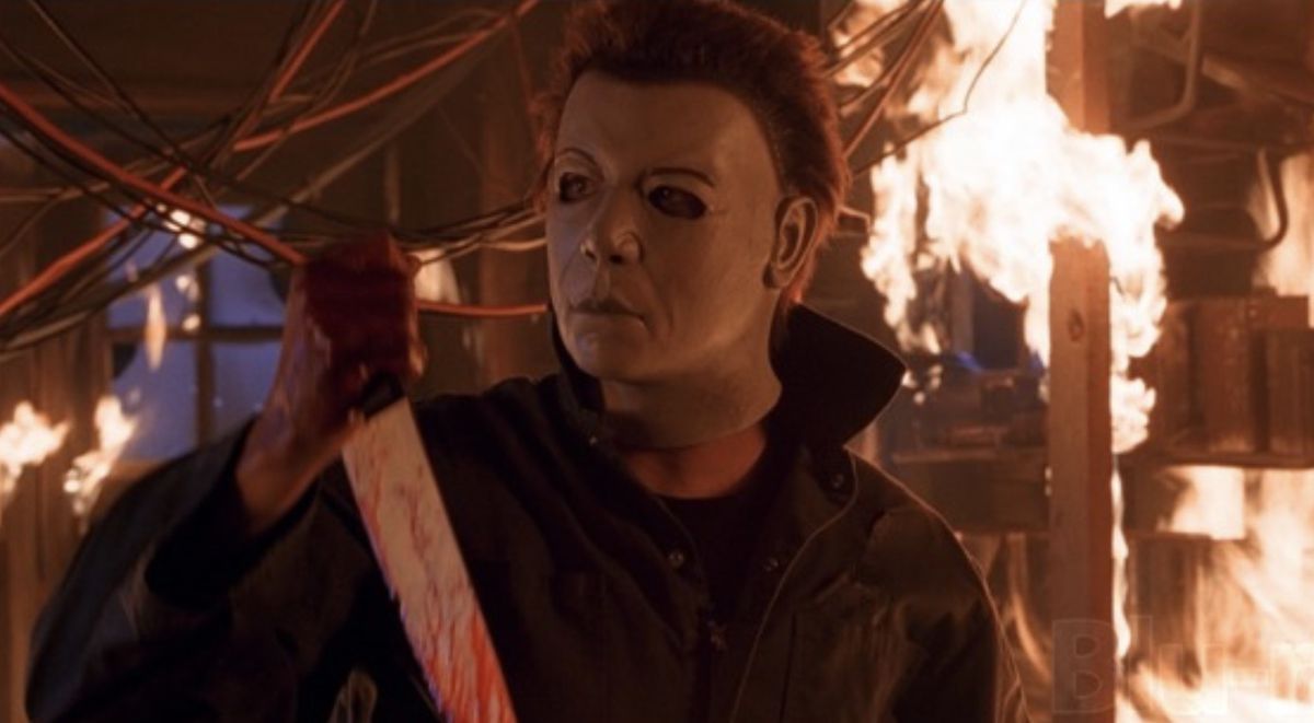 Michael Myers, surrounded by flames, wears a mask with raised eyebrows and a more pronounced nose.  He is holding a bloody knife.