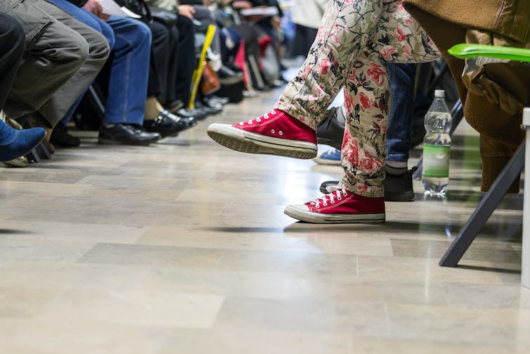 Feet of many people sit in a waiting room, with someone in red sneakers and floral jeans in the foreground and many in brown and black shoes in the background.