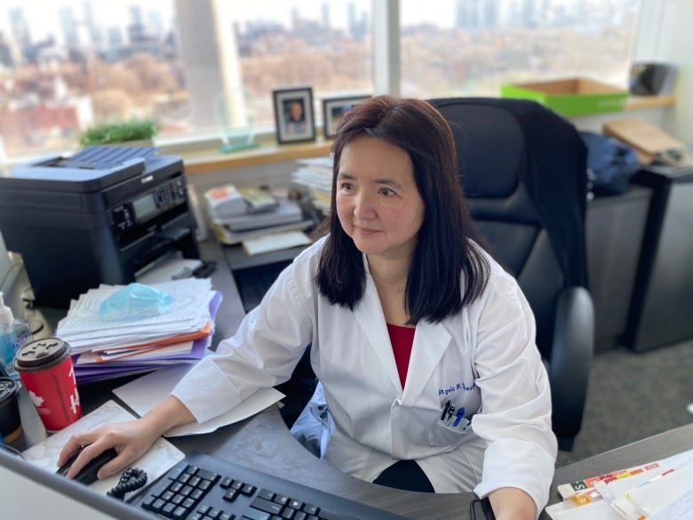 An Asian female doctor in a lab coat works on a computer in her office. 