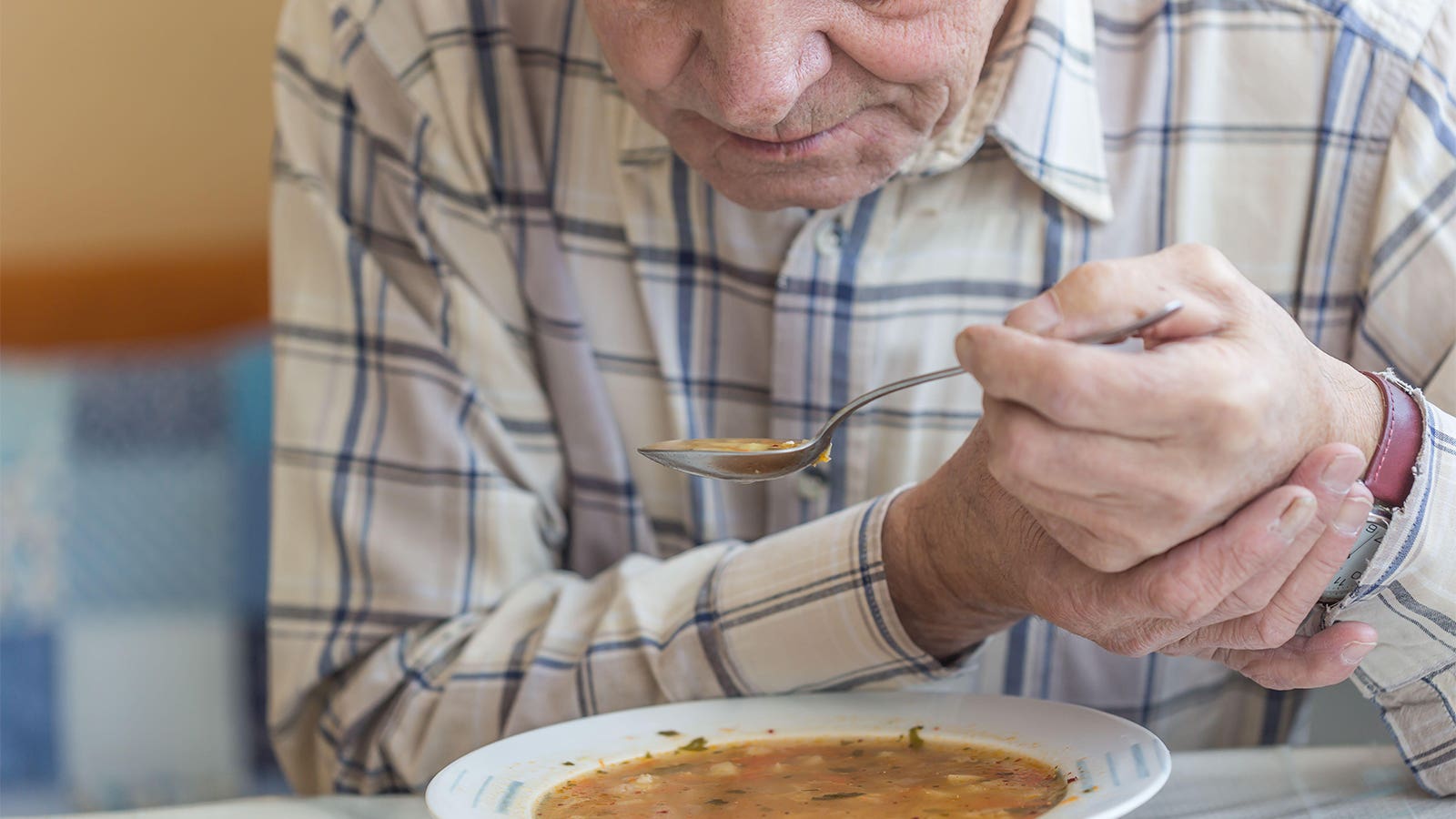 A photo of a senior man holding his steadying his hand which is holding a spoonful of soup.