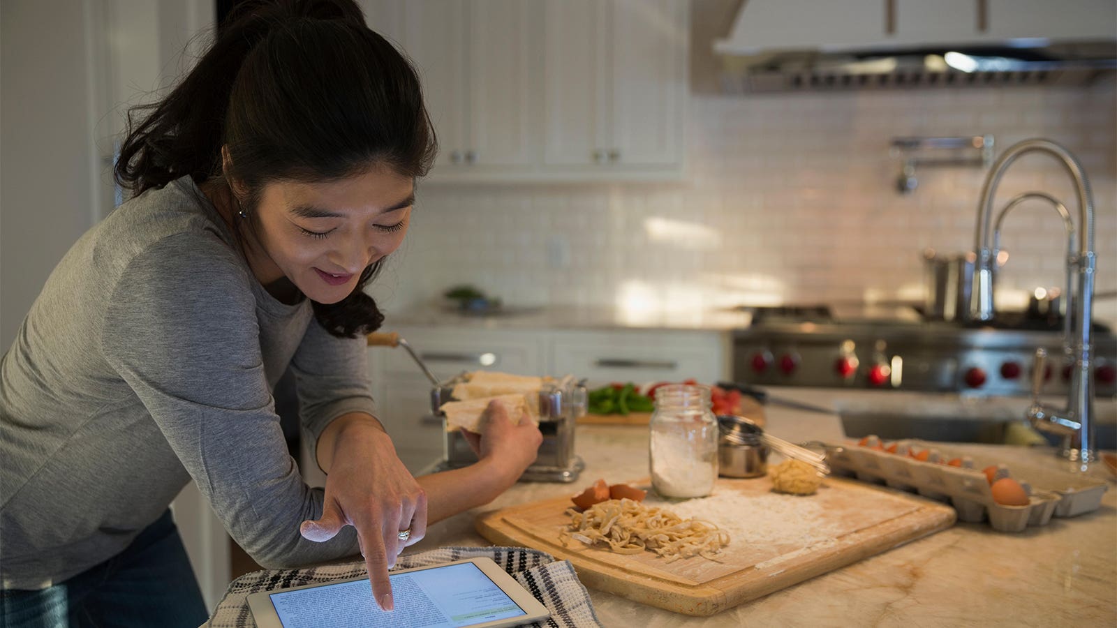 A photo of a woman consulting a tablet while using a pasta maker in her kitchen.