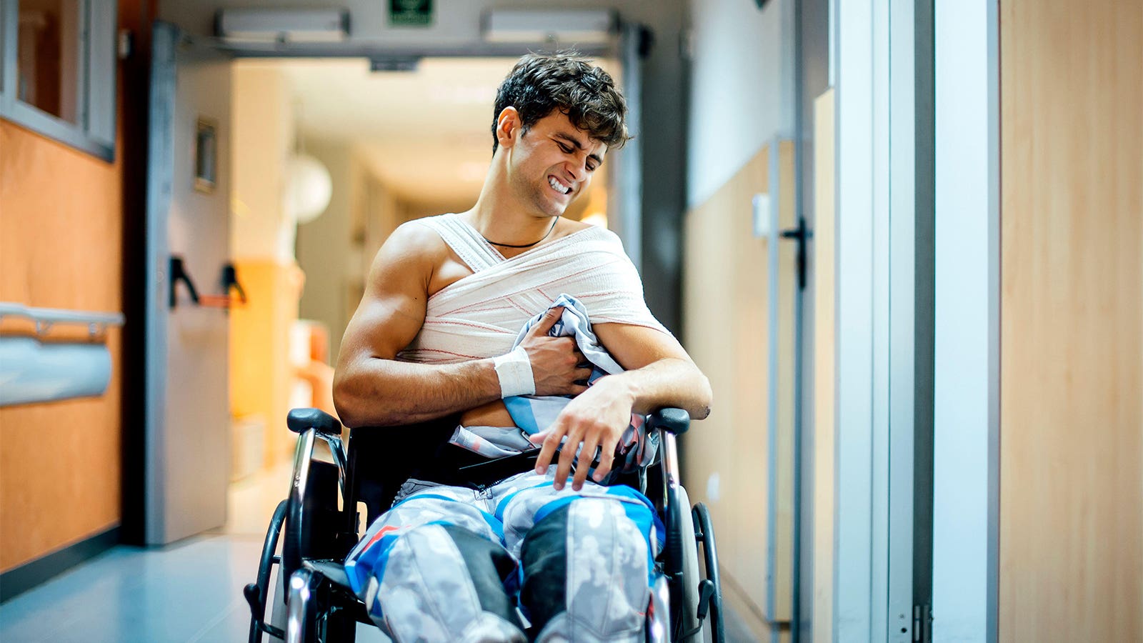 A photo of a man sitting in a wheelchair with his chest wrapped in bandages wincing in pain in a hospital.